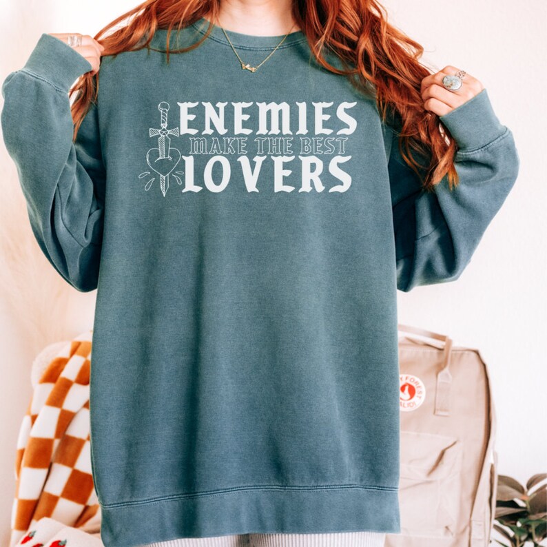 May include: A teal sweatshirt featuring the white text "ENEMIES MAKE THE BEST LOVERS". A sword and heart graphic is included. The top has long sleeves and a round neck.