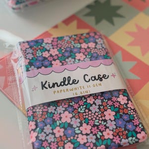 May include: Kindle Paperwhite II Gen (6.8in) case with a floral pattern in pink, blue, and white on a black background. The case is in a clear plastic package. The case is on a colorful patterned surface.