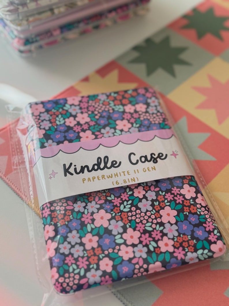 May include: Kindle Paperwhite II Gen (6.8in) case with a floral pattern in pink, blue, and white on a black background. The case is in a clear plastic package. The case is on a colorful patterned surface.