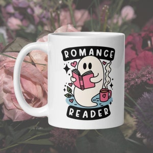 May include: White ceramic mug featuring a ghost reading a pink book and holding a cup of coffee. The mug has the text "Romance Reader" in black.