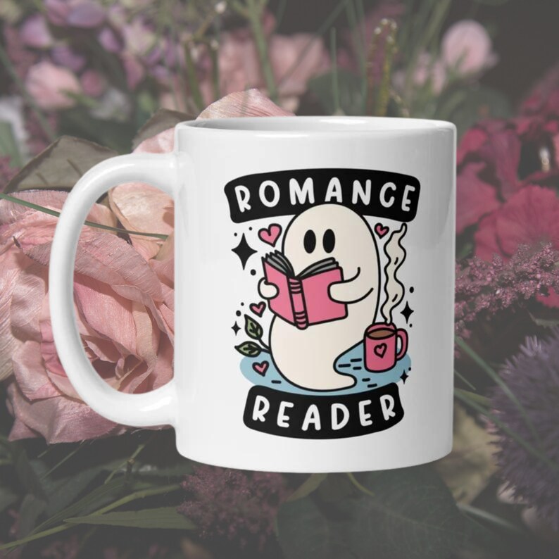 May include: White ceramic mug featuring a ghost reading a pink book and holding a cup of coffee. The mug has the text "Romance Reader" in black.