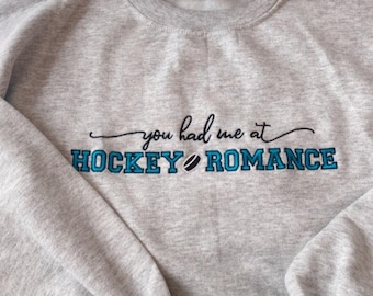 May include: A light gray sweatshirt with the phrase "you had me at HOCKEY ROMANCE" embroidered in black and teal. The word "hockey" features a hockey puck graphic. The sweatshirt is made of a soft material.