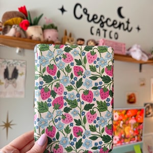 May include: A rectangular case with a cream background, featuring a pattern of pink strawberries, blue flowers, and green leaves. The case is held by a person. The text "Crescent CATCO" is visible in the background.