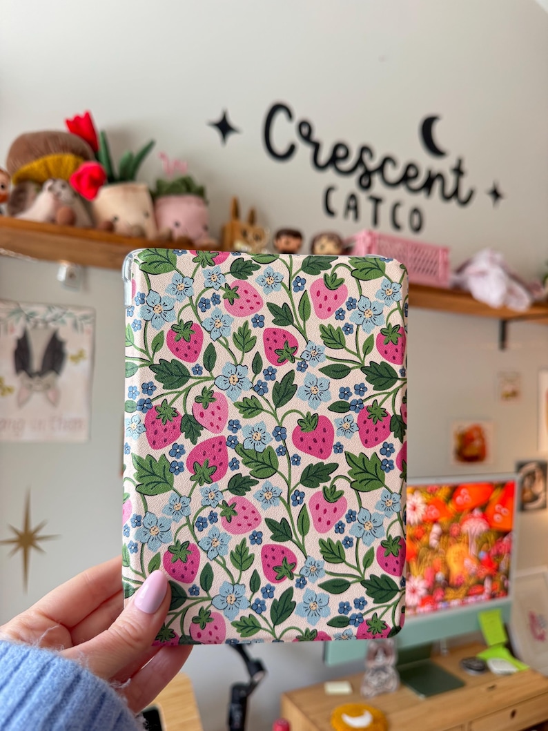 May include: A rectangular case with a cream background, featuring a pattern of pink strawberries, blue flowers, and green leaves. The case is held by a person. The text "Crescent CATCO" is visible in the background.