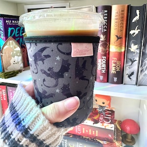 Peut inclure: Un manchon de tasse à café gris foncé avec un motif de dragon et d'étoiles, tenu par une personne portant un pull crème. La tasse contient une boisson et est surmontée d'un couvercle en plastique transparent. Le manchon porte une étiquette rose avec l'inscription "Crescent Cakes".