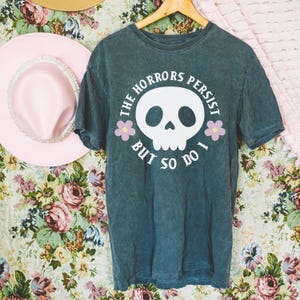 May include: Dark teal t-shirt with a white skull graphic and the text "THE HORRORS PERSIST BUT SO DO I." The design includes two pink flowers. A pink hat with a pearl band is visible in the background.
