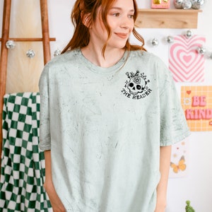 May include: A light green t-shirt with a distressed look featuring a black skull with flowers and the text "Only the reader" printed on the front.