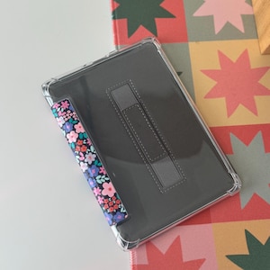 May include: A clear tablet case with a floral patterned stylus holder and a gray stand. The floral pattern includes pink, blue, and white flowers on a black background. The tablet case is on a colorful star-patterned surface.