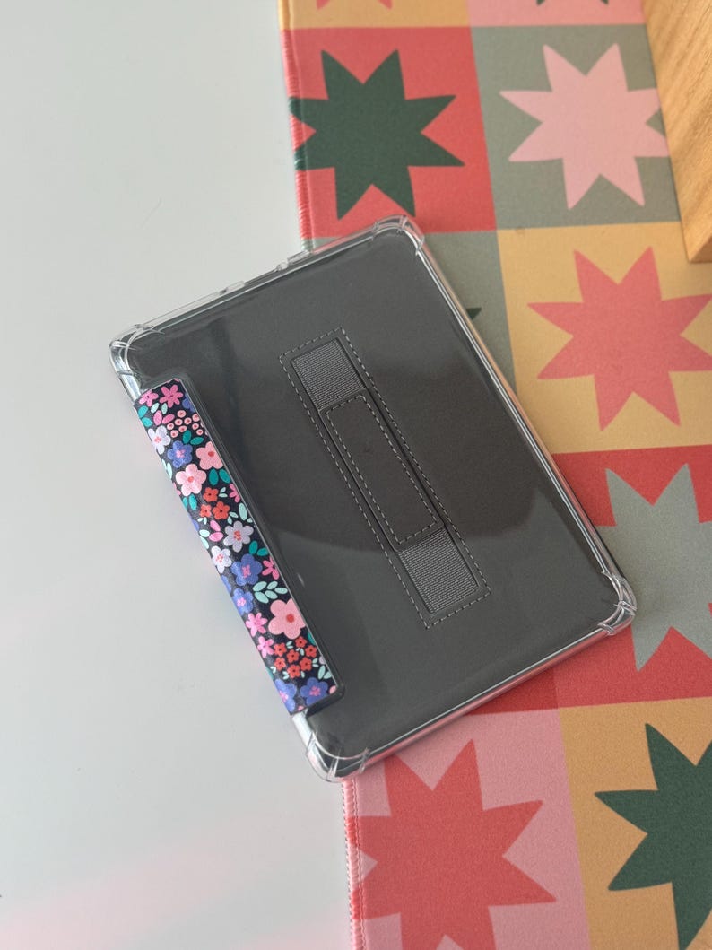 May include: A clear tablet case with a floral patterned stylus holder and a gray stand. The floral pattern includes pink, blue, and white flowers on a black background. The tablet case is on a colorful star-patterned surface.