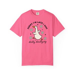 May include: Pink t-shirt with a cartoon duck graphic. The duck is white with an orange beak and feet, surrounded by daisies and the text "Sorry I'm late I was Dilly Dallying."