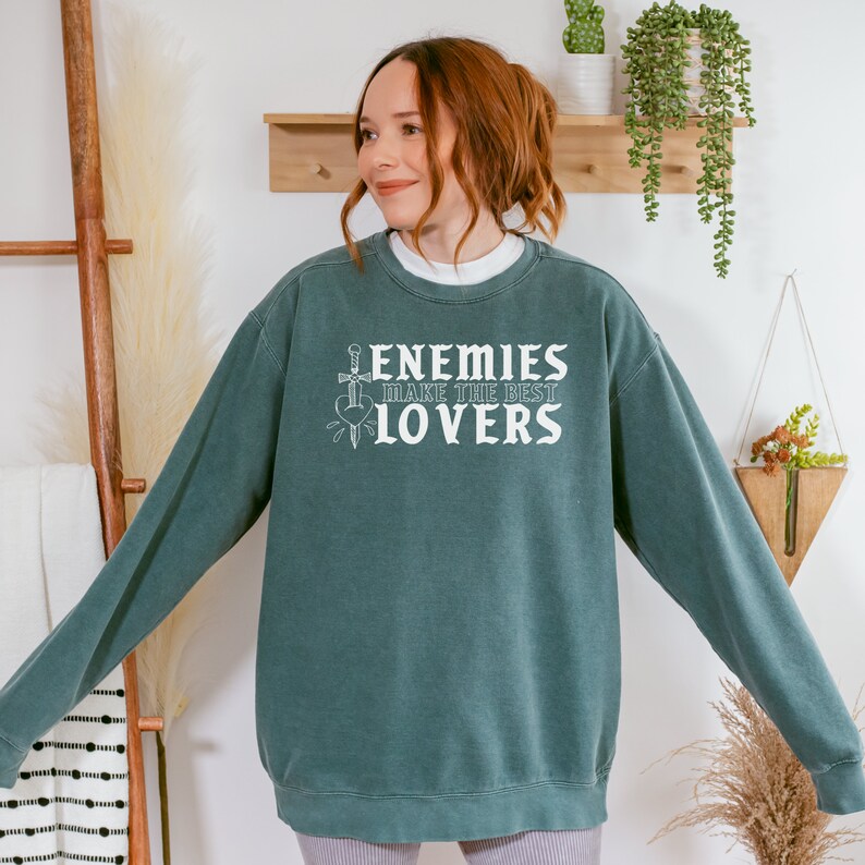 May include: A teal sweatshirt with the text "ENEMIES MAKE THE BEST LOVERS" in white. The graphic includes a sword piercing a heart. The sweatshirt is a crew neck style.
