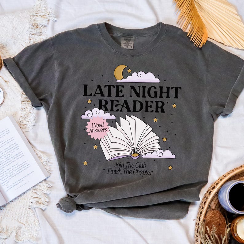 May include: Dark gray t-shirt with the text "LATE NIGHT READER" and an illustration of an open book, moon, stars, and clouds. The shirt also includes the phrases "I Need Answers", "Join The Club", and "Finish The Chapter".