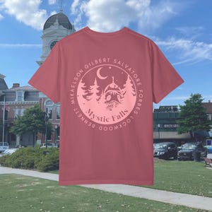 Puede incluir: Una camiseta roja con un gr&aacute;fico circular rosa en la espalda. El gr&aacute;fico presenta una luna creciente, estrellas, &aacute;rboles y una casa con el texto "Mystic Falls" y los nombres de los personajes principales del programa de televisi&oacute;n "The Vampire Diaries".
