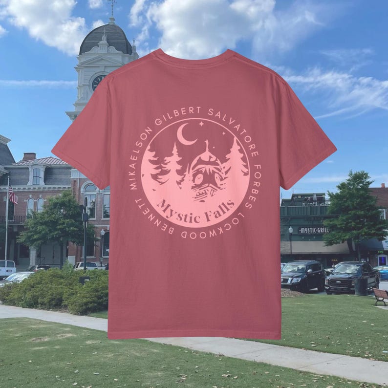 Puede incluir: Una camiseta roja con un gr&aacute;fico circular rosa en la espalda. El gr&aacute;fico presenta una luna creciente, estrellas, &aacute;rboles y una casa con el texto "Mystic Falls" y los nombres de los personajes principales del programa de televisi&oacute;n "The Vampire Diaries".