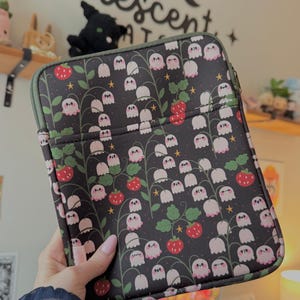 May include: A rectangular black tablet sleeve featuring a repeating pattern of white lily of the valley flowers with pink ghost-like faces, red strawberries, green leaves, and small yellow stars. The sleeve has a dark green zipper and trim.