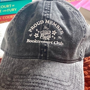 May include: A black denim baseball cap with the text "Proud Member of the Booktrovert Club" embroidered in white on the front. The embroidery includes a small illustration of a building with stars.