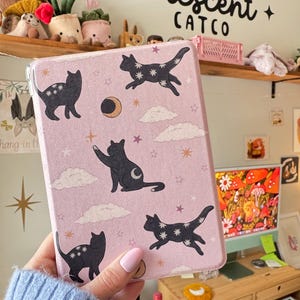 May include: A pink tablet case featuring black cat illustrations with star patterns, moons, and clouds. The case has the text "Crescent Catco" in black. The case is held in a person's hand.