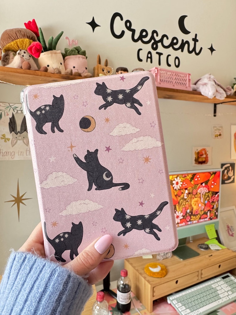 May include: A pink tablet case featuring black cat illustrations with star patterns, moons, and clouds. The case has the text "Crescent Catco" in black. The case is held in a person's hand.