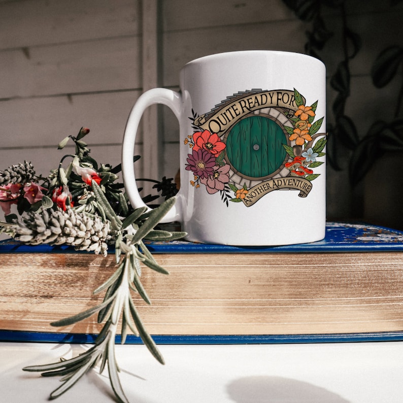 May include: White ceramic mug with a green door design and floral accents. The mug reads "Quite Ready For Another Adventure".