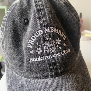 May include: A black denim baseball cap with white embroidered text that reads "Proud Member of the Booktrovert Club".