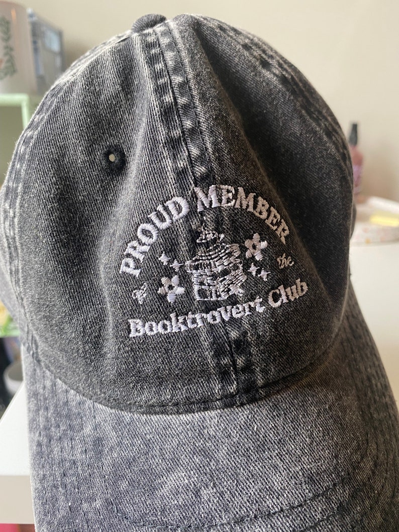 May include: A black denim baseball cap with white embroidered text that reads "Proud Member of the Booktrovert Club".
