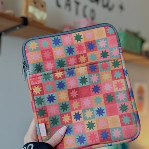 May include: A colorful tablet sleeve with a star pattern in a patchwork design. The sleeve features a zipper closure and a front pocket. The color palette includes red, pink, orange, yellow, blue, and green.