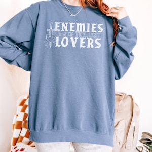 May include: A pale blue sweatshirt featuring the white text "ENEMIES MAKE THE BEST LOVERS". The design includes a sword through a heart. The sweatshirt has a crew neck.