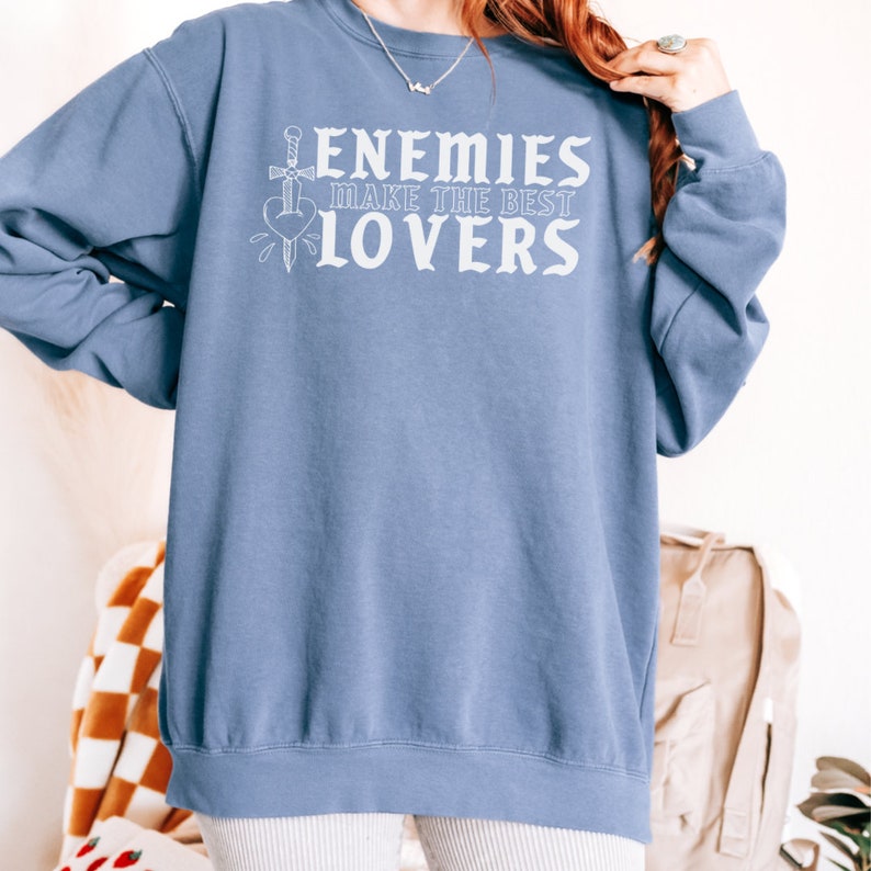 May include: A pale blue sweatshirt featuring the white text "ENEMIES MAKE THE BEST LOVERS". The design includes a sword through a heart. The sweatshirt has a crew neck.