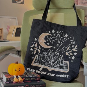 Puede incluir: Bolso tote negro con un dise&ntilde;o espeluznante que presenta una luna creciente, estrellas, murci&eacute;lagos y el texto "READ BOOKS STAY SPOOKY". La bolsa est&aacute; colgada en una silla verde. Una peque&ntilde;a calabaza y libros est&aacute;n frente a la bolsa.