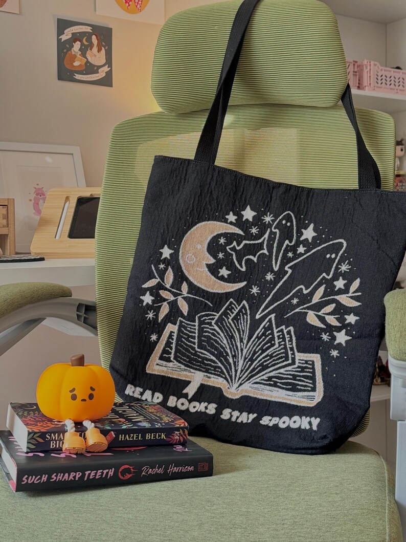 Puede incluir: Bolso tote negro con un dise&ntilde;o espeluznante que presenta una luna creciente, estrellas, murci&eacute;lagos y el texto "READ BOOKS STAY SPOOKY". La bolsa est&aacute; colgada en una silla verde. Una peque&ntilde;a calabaza y libros est&aacute;n frente a la bolsa.