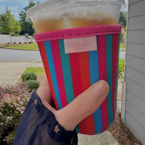 May include: A clear plastic cup filled with an iced beverage, held in a colorful striped neoprene sleeve. The sleeve features vertical stripes in red, blue, teal, and pink, with a pink top and a small label that reads "Crescent Cafe".