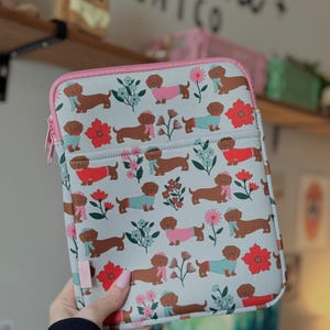 May include: A light gray tablet case with a pink zipper and trim. The case features a repeating pattern of brown dachshunds wearing hats and scarves, interspersed with red and pink flowers and blue floral sprigs.