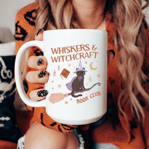 May include: White ceramic mug with the text "WHISKERS & WITCHCRAFT BOOK CLUB". The mug features a black cat wearing a witch hat and cape, riding a broom. Additional illustrations include a book, candles, stars, and a crescent moon.
