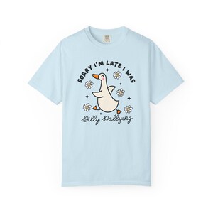 May include: Light blue t-shirt with a cartoon goose illustration. The goose is white with an orange beak and feet, surrounded by daisies and the text "Sorry I'm late I was dilly dallying."