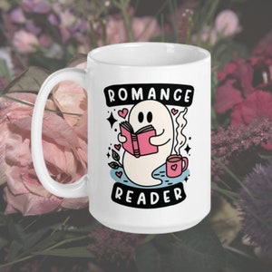 May include: White ceramic mug featuring a ghost reading a pink book and holding a cup of coffee. The mug is inscribed with the text "Romance Reader" in black lettering.