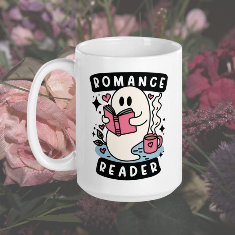 May include: White ceramic mug featuring a ghost reading a pink book and holding a cup of coffee. The mug is inscribed with the text "Romance Reader" in black lettering.