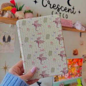 May include: A rectangular, patterned case featuring a repeating design of purple dragons, bows, flowers, and butterflies on a light green background. The case is held in front of a white wall with the text "Crescent Cat Co."