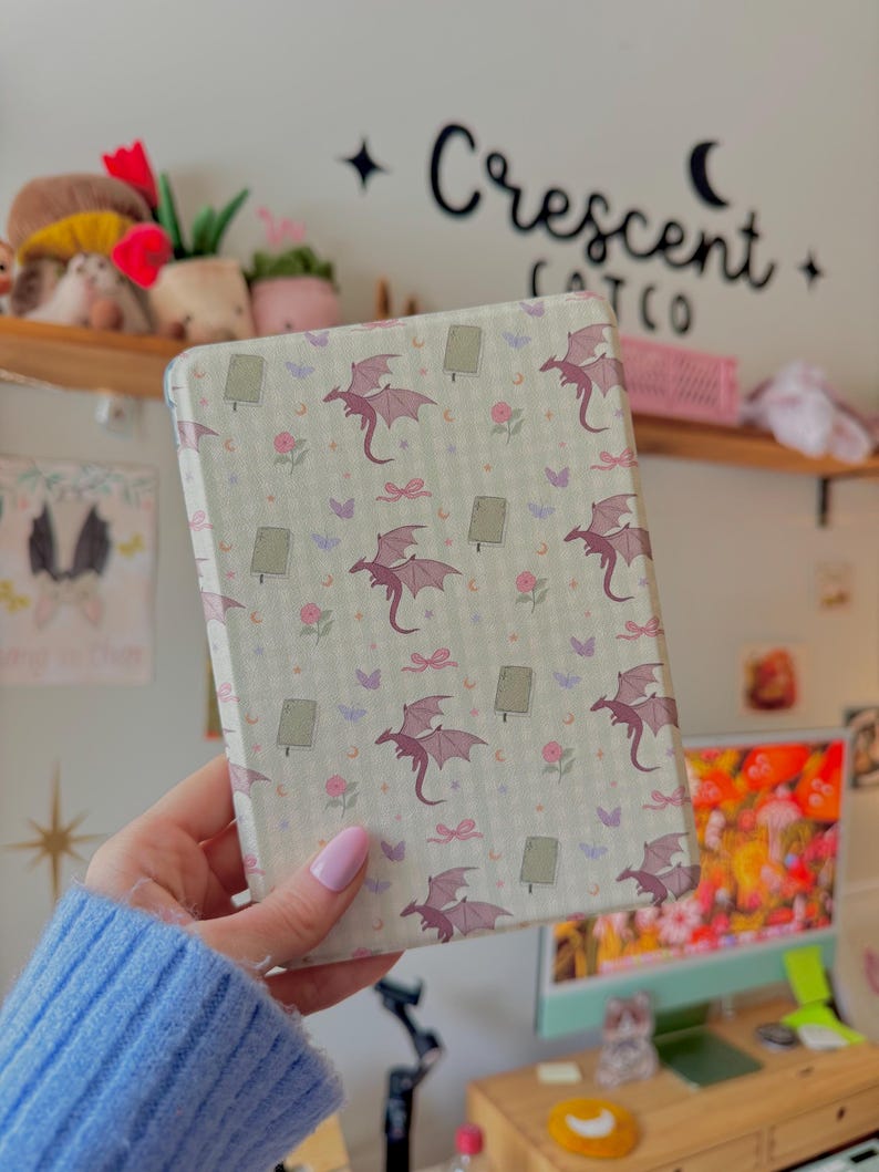 May include: A rectangular, patterned case featuring a repeating design of purple dragons, bows, flowers, and butterflies on a light green background. The case is held in front of a white wall with the text "Crescent Cat Co."