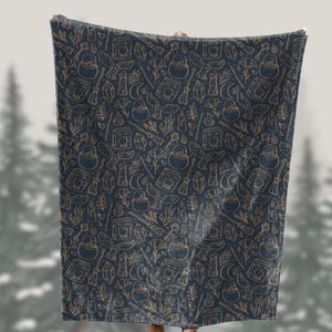 May include: A navy blue blanket with a repeating pattern of gold-colored witchy elements, including cauldrons, books, crystals, brooms, and moons. The blanket is held up, showcasing the full design and soft texture. The overall theme is mystical and magical.