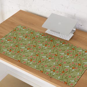 May include: A rectangular desk mat with a green background featuring a repeating pattern of ladybugs, pink flowers, and white flowers with green leaves. The mat is on a wooden desk.