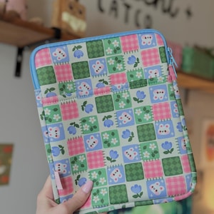 May include: A colorful tablet sleeve featuring a patchwork design with squares of pink gingham, green, and light green. The squares are decorated with white flowers, blue flowers, and bear and butterfly graphics. The sleeve has a light blue zipper.