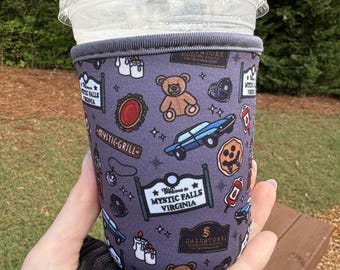 May include: A purple drinks sleeve featuring various themed illustrations, such as a teddy bear, car, and the text "Welcome to Mystic Falls Virginia." The sleeve is designed to fit a cold beverage cup.