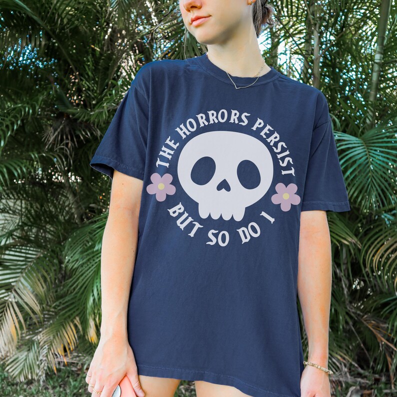 May include: Navy blue t-shirt featuring a white skull design and the text "THE HORRORS PERSIST BUT SO DO I." The design includes two pink flower accents. A comfortable, casual style.