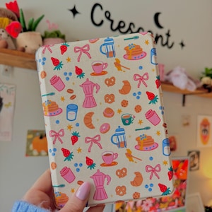 May include: A cream-colored tablet case featuring a repeating pattern of breakfast-themed illustrations. The design includes coffee cups, strawberries, pancakes, croissants, and bows. The word "Crescent" is written in black script on the wall behind the case.