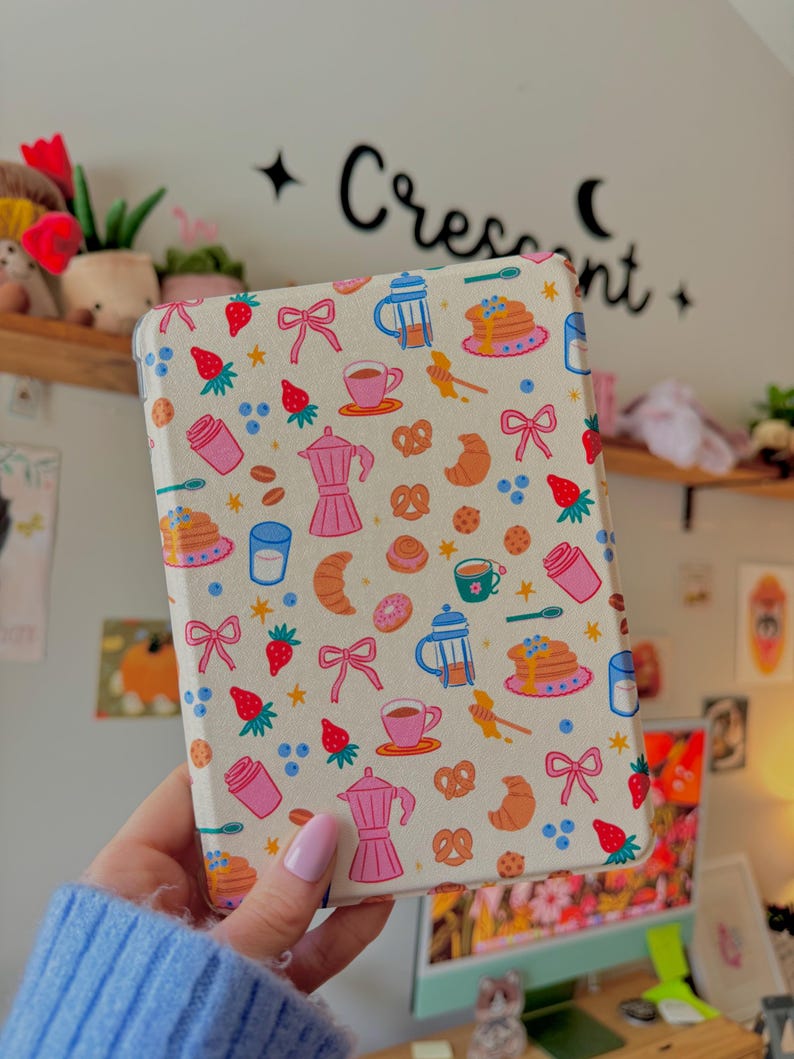 May include: A cream-colored tablet case featuring a repeating pattern of breakfast-themed illustrations. The design includes coffee cups, strawberries, pancakes, croissants, and bows. The word "Crescent" is written in black script on the wall behind the case.