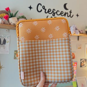 Peut inclure: Housse de tablette avec un motif floral et vichy. La partie supérieure est orange avec des fleurs de marguerite blanches, et la partie inférieure est vichy orange et blanc. Elle a une fermeture éclair jaune et une petite étiquette rose.