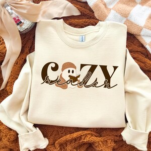 May include: Cream-coloured sweatshirt featuring the text "Cozy Reader" in brown script, with a graphic of a ghost wearing a beanie and reading a book. The sweatshirt has long sleeves and a crew neck.