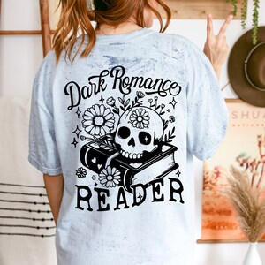 May include: A light blue t-shirt with a distressed look featuring a black and white graphic of a skull with flowers and a book. The text "Dark Romance Reader" is printed on the shirt.