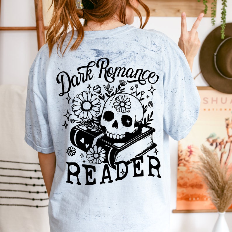 May include: A light blue t-shirt with a distressed look featuring a black and white graphic of a skull with flowers and a book. The text "Dark Romance Reader" is printed on the shirt.