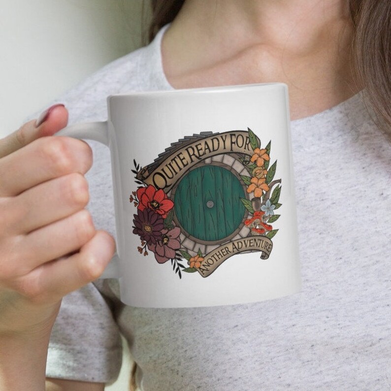 May include: White ceramic mug with a green door and floral design. The text on the mug reads "Quite ready for another adventure".